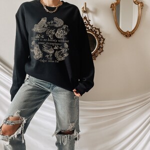 May include: Black sweatshirt with a graphic print of frogs and mushrooms. The text "There is no beauty without some strangeness - Edgar Allan Poe" is printed on the front of the sweatshirt.