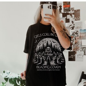 Może przedstawiać: Czarny t-shirt z grafiką przedstawiającą czaszkę, las i postacie w pelerynach. Na koszulce nadrukowano napisy "GIRLS COILL BE" i "READING COVEN". Na koszulce wydrukowano r&oacute;wnież frazę "BURNING STEREOTYPES NOT BOOKS".
