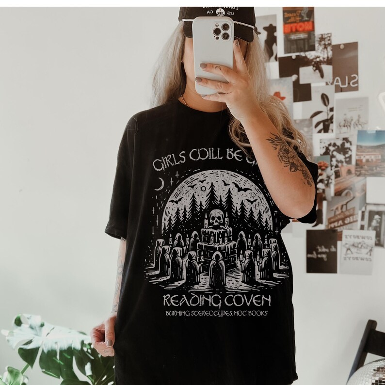 Może przedstawiać: Czarny t-shirt z grafiką przedstawiającą czaszkę, las i postacie w pelerynach. Na koszulce nadrukowano napisy "GIRLS COILL BE" i "READING COVEN". Na koszulce wydrukowano r&oacute;wnież frazę "BURNING STEREOTYPES NOT BOOKS".