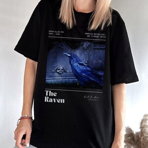 May include: Black t-shirt with a graphic print of a raven perched on a tombstone. The text "The Raven" is printed below the image. The text "Edgar Allan Poe 1809 - 1849 American Writer, Poet, and Literary Critic" is printed above the image.