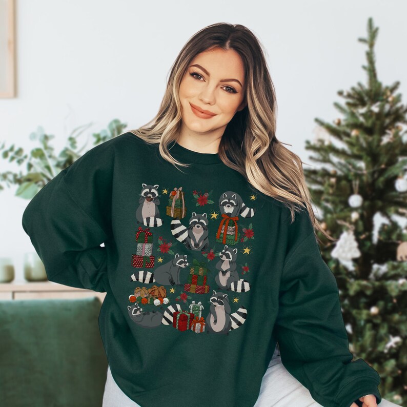 K&ouml;nnte beinhalten: Ein dunkelgr&uuml;ner Pullover mit einem weihnachtlichen Design, das Waschb&auml;ren zeigt, die Geschenke halten und von Sternen, Blumen und Ornamenten umgeben sind.