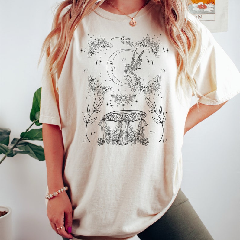Pu&ograve; includere: Una maglietta color crema con un design grafico in bianco e nero di una fata seduta su una luna crescente con un fungo e farfalle. Il design include anche stelle e foglie.
