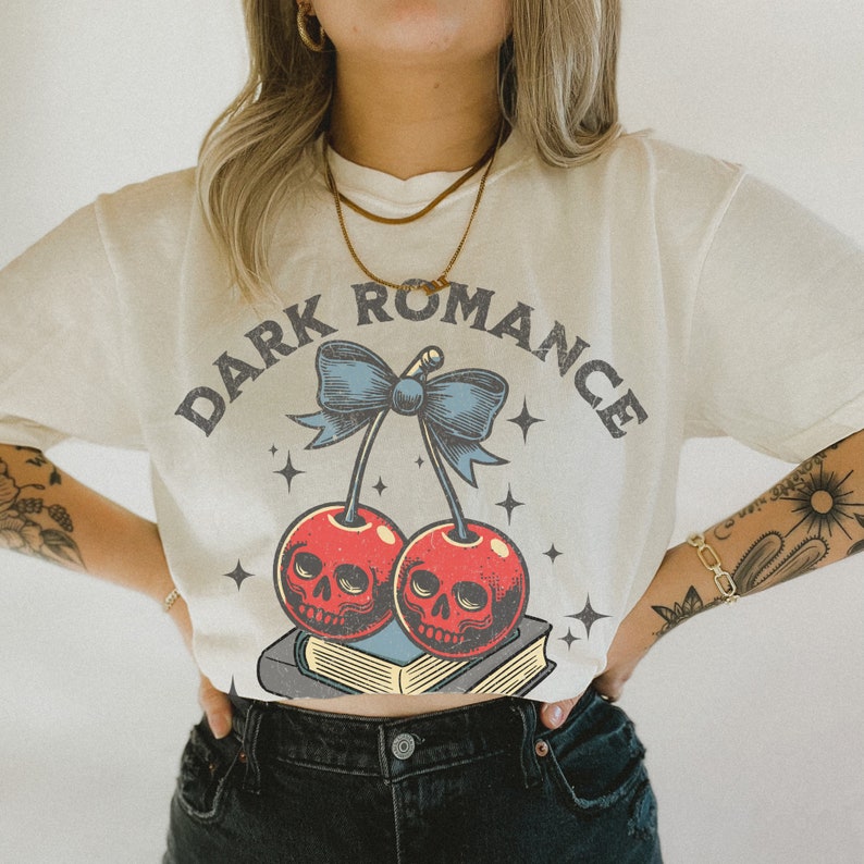K&ouml;nnte beinhalten: Cremefarbenes T-Shirt mit dem Text "DARK ROMANCE" &uuml;ber einer Grafik von zwei Kirschen mit Totenkopfgesichtern, einer blauen Schleife und Sternen. Die Kirschen liegen auf einem Stapel B&uuml;cher. Das Shirt hat einen Vintage-Look.