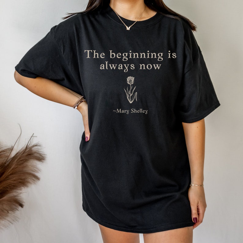 May include: Black oversized t-shirt with the quote "The beginning is always now" and a small flower graphic. The quote is attributed to Mary Shelley.