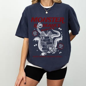 Pu&ograve; includere: T-shirt blu navy con la scritta rossa "MONSTER ROMANCE BOOK CLUB". Il design presenta una pila di libri con un polpo e cuori. La maglietta ha anche le frasi "ONE BED, SOLOS, NO REGRETS, MEETS NIGHTLY" e "COME FOR THE TENTACLES".