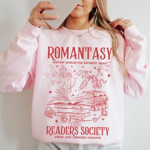 May include: Pink sweatshirt with a red graphic design that says "Romantasy: Fantasy Worlds for Romantic Souls" and "Readers Society: Where Love Conquers Kingdoms". The graphic features a winged cupid, a dragon, a stack of books, a key, a padlock, and roses.