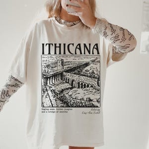 May include: A white t-shirt with a black graphic print of a bridge and the text "ITHICANA". The text below the graphic reads "Raging seas, hidden jungles and a bridge of secrets" and "Roled by King Arun Kedell".