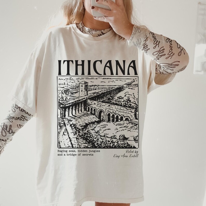 May include: A white t-shirt with a black graphic print of a bridge and the text "ITHICANA". The text below the graphic reads "Raging seas, hidden jungles and a bridge of secrets" and "Roled by King Arun Kedell".