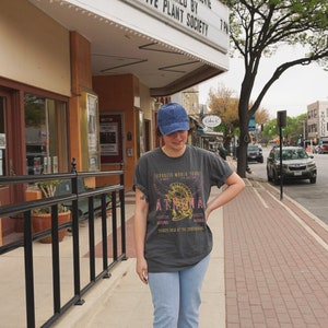 Puede incluir: Una persona lleva una camiseta gris oscura con un gr&aacute;fico de un casco y el texto "Goddess World Tour" y "Athena". La camiseta tambi&eacute;n tiene el texto "Special Guests" y "Tickets sold at the crossroads". La persona lleva una gorra de mezclilla azul y jeans azules.