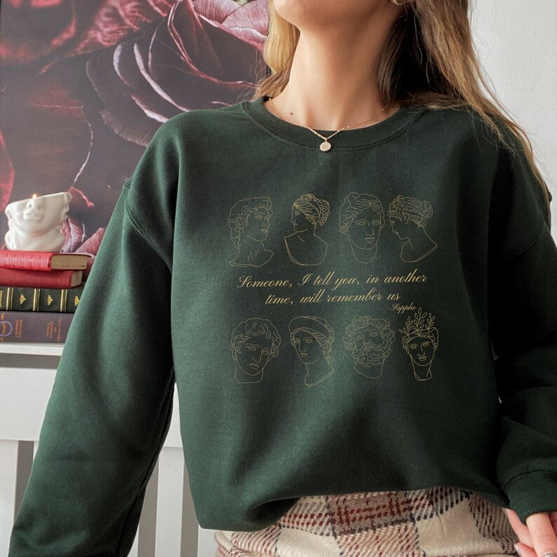 Pode incluir: Uma camisola verde escura com um contorno dourado de v&aacute;rios rostos e o texto "Algu&eacute;m, eu te digo, em outro tempo, se lembrar&aacute; de n&oacute;s - Safo".