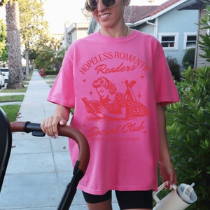 Peut inclure: T-shirt rose avec un graphique rouge d'une femme lisant un livre. Le graphique dit "Hopeless Romantic Readers Social Club. Home of the Bookworm Burn."