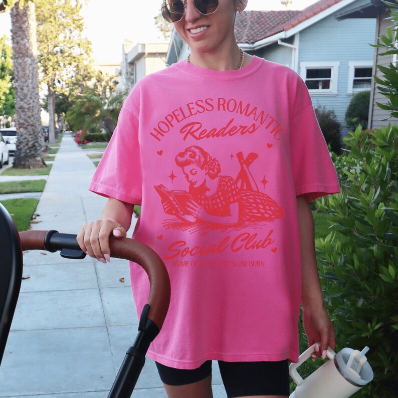 Peut inclure: T-shirt rose avec un graphique rouge d'une femme lisant un livre. Le graphique dit "Hopeless Romantic Readers Social Club. Home of the Bookworm Burn."