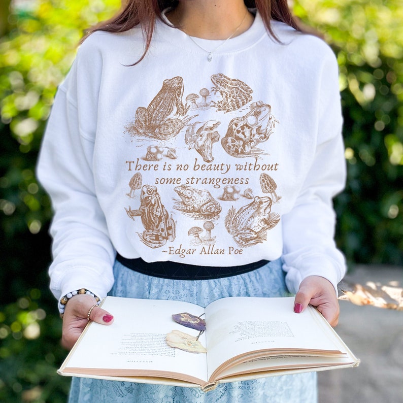 May include: White sweatshirt with a brown illustration of frogs and mushrooms. The text "There is no beauty without some strangeness - Edgar Allan Poe" is printed on the sweatshirt.