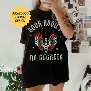 May include: Black t-shirt with the text "BOOK ADDICT" above a colorful moth design and the words "NO REGRETS" below. The moth has teal, red, and yellow accents. A yellow starburst graphic with the text "LUCABOOCO ORIGINAL DESIGN" is in the upper left.