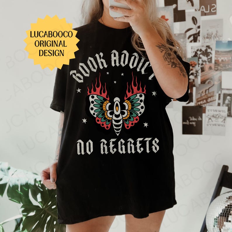 May include: Black t-shirt with the text "BOOK ADDICT" above a colorful moth design and the words "NO REGRETS" below. The moth has teal, red, and yellow accents. A yellow starburst graphic with the text "LUCABOOCO ORIGINAL DESIGN" is in the upper left.