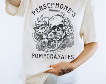 May include: A white t-shirt with a black graphic design of a skull surrounded by pomegranates and flowers. The text "Persephone's Farm Fresh Pomegranates" is printed on the shirt.