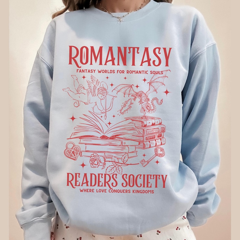 May include: Light blue sweatshirt with a red graphic design featuring a winged cherub, a dragon, and a stack of books. The text reads "Romantasy: Fantasy Worlds for Romantic Souls" and "Readers Society: Where Love Conquers Kingdoms".