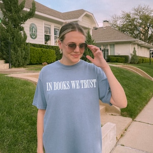 May include: A woman wearing sunglasses and a light blue t-shirt with the text "In Books We Trust" printed in white.