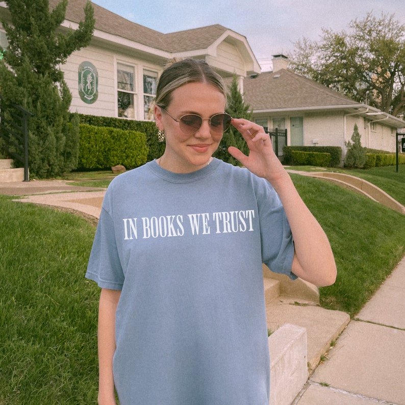 May include: A woman wearing sunglasses and a light blue t-shirt with the text "In Books We Trust" printed in white.