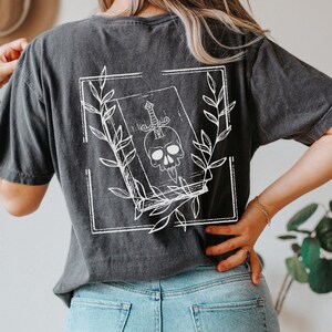 May include: A black t-shirt with a white graphic design of a book with a skull and a dagger inside. The book is surrounded by leaves and is framed in a square.