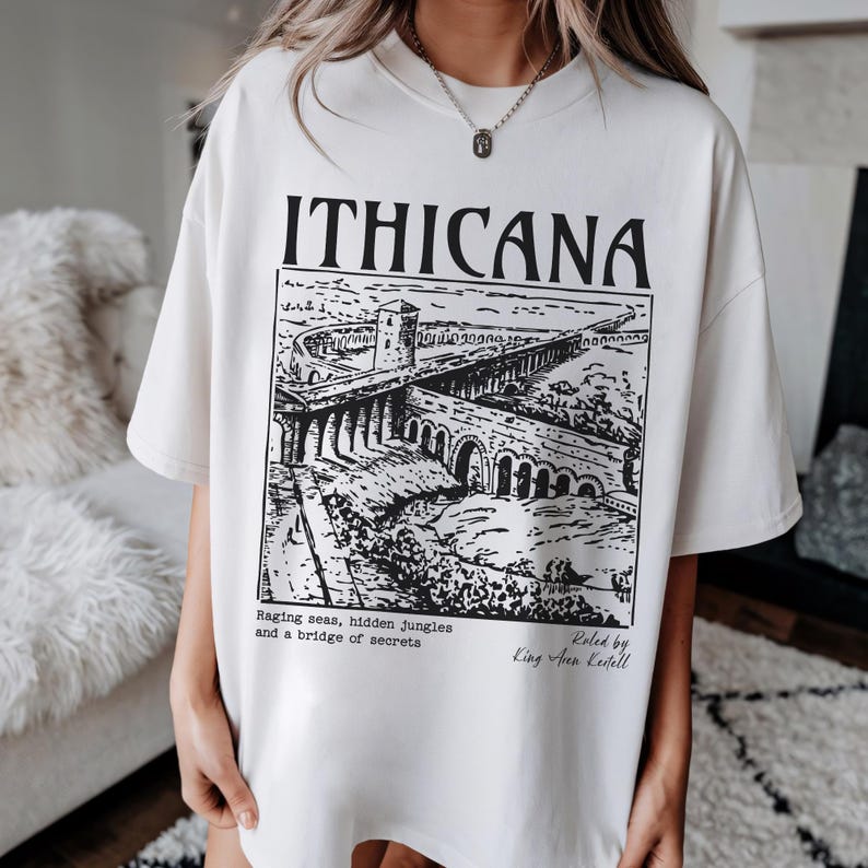 May include: White oversized t-shirt with a black and white graphic print of a bridge and a castle. The text "ITHICANA" is printed in large letters at the top of the graphic. The text "Raging seas, hidden jungles and a bridge of secrets" is printed below the graphic. The text "Roled by King Aren Kenell" is printed in the bottom right corner of the graphic.