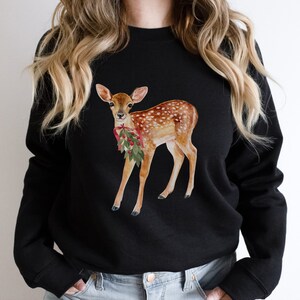 May include: Black sweatshirt with a watercolor illustration of a fawn with a red and green holly berry sprig in its mouth.