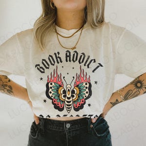 May include: Cream-colored t-shirt with the words "BOOK ADDICT" in a gothic font above a colorful butterfly design. The butterfly has teal, pink, and yellow accents with flames. Small stars surround the butterfly.