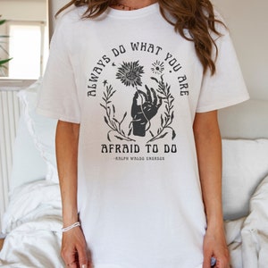 以下が含まれることがあります： 白いTシャツに、黒い花の絵が描かれた手のグラフィックプリントが施されています。テキストには「Always do what you are afraid to do - Ralph Waldo Emerson」と書いてあります。