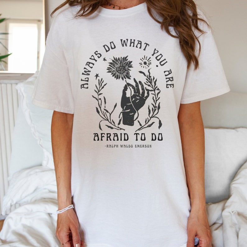 以下が含まれることがあります： 白いTシャツに、黒い花の絵が描かれた手のグラフィックプリントが施されています。テキストには「Always do what you are afraid to do - Ralph Waldo Emerson」と書いてあります。