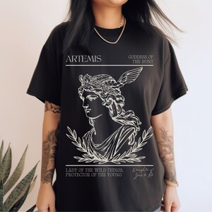 May include: Black t-shirt with a white graphic of Artemis, the Greek goddess of the hunt. The graphic includes a line drawing of Artemis with a crown and a bow and arrow. The text "ARTEMIS", "GODDESS OF THE HUNT", "LADY OF THE WILD THINGS. PROTECTOR OF THE YOUNG", and "Daughter of Zeus & Leto" is included in the graphic.