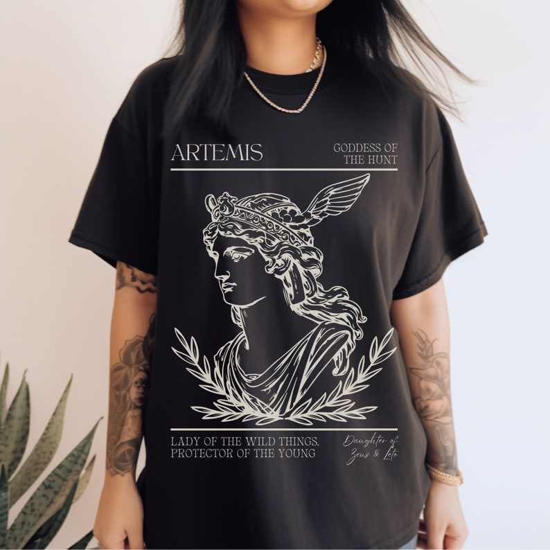 May include: Black t-shirt with a white graphic of Artemis, the Greek goddess of the hunt. The graphic includes a line drawing of Artemis with a crown and a bow and arrow. The text "ARTEMIS", "GODDESS OF THE HUNT", "LADY OF THE WILD THINGS. PROTECTOR OF THE YOUNG", and "Daughter of Zeus & Leto" is included in the graphic.