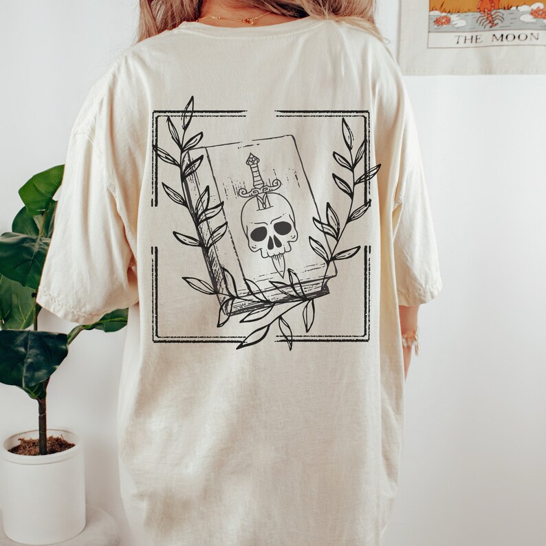May include: A cream colored t-shirt with a black and white graphic of a book with a skull and a dagger inside. The book is surrounded by leaves and is framed in a black square.