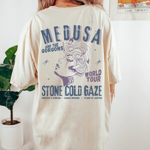 May include: A beige t-shirt with a graphic print of Medusa, a mythical Greek figure with snakes for hair. The text "MEDUSA AND THE GORGONS WORLD TOUR STONE COLD GAZE SERPENT'S SCREAM - HADES INFERNO - STARE OF JUSTICE" is printed below the image.