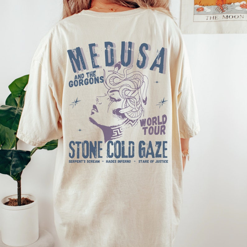 May include: A beige t-shirt with a graphic print of Medusa, a mythical Greek figure with snakes for hair. The text "MEDUSA AND THE GORGONS WORLD TOUR STONE COLD GAZE SERPENT'S SCREAM - HADES INFERNO - STARE OF JUSTICE" is printed below the image.