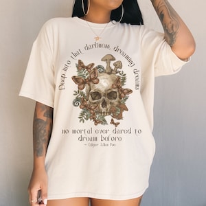 Pu&ograve; includere: Una maglietta color crema con un teschio circondato da funghi, farfalle e foglie. Il testo recita "Deep into that darkness, dreaming dreams no mortal ever dared to dream before - Edgar Allan Poe".