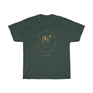 May include: A dark green t-shirt with a gold circle design featuring a moon, sun, and hands holding a branch with leaves. The text inside the circle reads "I took my power in my hand and went against the world." - Emily Dickinson.