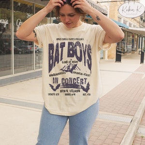 K&ouml;nnte beinhalten: Ein beiges T-Shirt mit einem Vintage-Konzertposter-Design mit dem Text "Bat Boys" in einer verwitterten Schriftart. Das Poster enth&auml;lt auch den Text "Lord of Night", "Shadowsinger", "Warrior-Heart", "In Concert", "Rita's Velaris", "Tickets 20&euro;", "Doors 8PM" und "Oct. 4th".