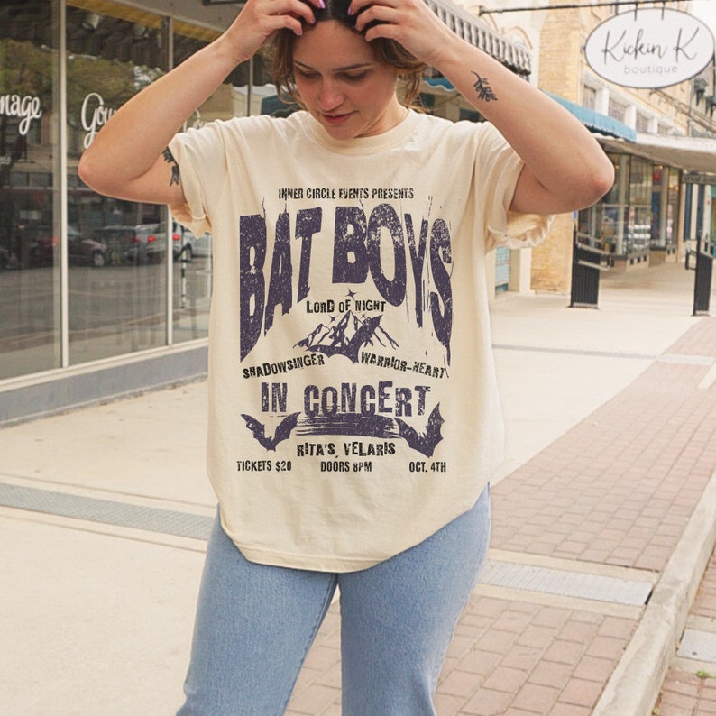 K&ouml;nnte beinhalten: Ein beiges T-Shirt mit einem Vintage-Konzertposter-Design mit dem Text "Bat Boys" in einer verwitterten Schriftart. Das Poster enth&auml;lt auch den Text "Lord of Night", "Shadowsinger", "Warrior-Heart", "In Concert", "Rita's Velaris", "Tickets 20&euro;", "Doors 8PM" und "Oct. 4th".