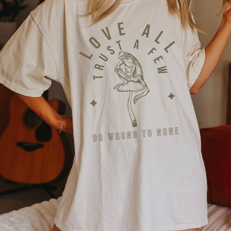 May include: White t-shirt with a brown frog sitting on a mushroom. The shirt has the text "Love All Trust A Few Do Wrong To None" printed on it.