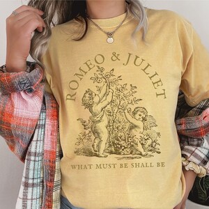 May include: A yellow t-shirt with a vintage illustration of two cherubs holding flowers. The text "ROMEO & JULIET" is in a circle around the cherubs. The text "WHAT MUST BE SHALL BE" is below the illustration.