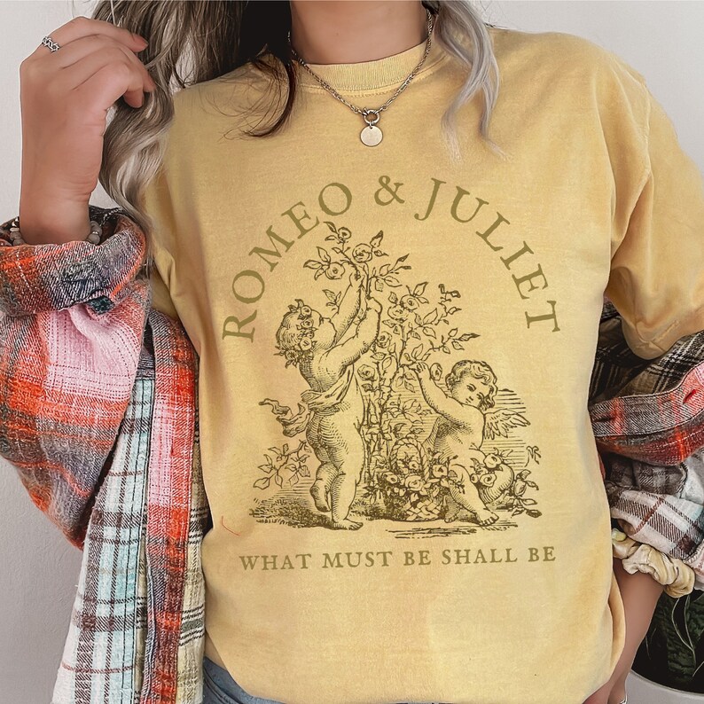 May include: A yellow t-shirt with a vintage illustration of two cherubs holding flowers. The text "ROMEO & JULIET" is in a circle around the cherubs. The text "WHAT MUST BE SHALL BE" is below the illustration.