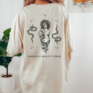 May include: A cream colored t-shirt with a black and white graphic print of a woman with snakes and the text "STRENGTH BEAUTY POWER".