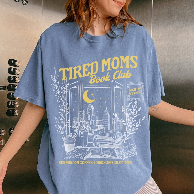 May include: A light blue t-shirt with a white graphic design of a cityscape at night with a crescent moon and stars. The text "TIRED MOMS Book Club" is in yellow lettering above the graphic. The text "NIGHTLY AT 10PM" is in a white rectangle below the graphic. The text "RUNNING ON COFFEE, CHAOS AND CHAPTERS" is in white lettering below the graphic.