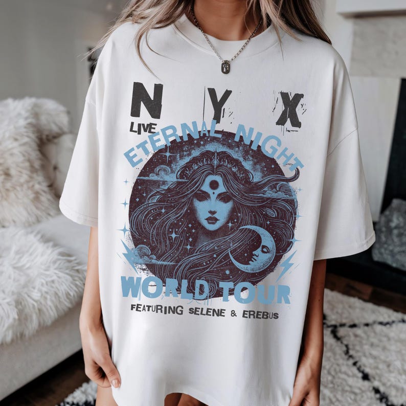 May include: Off-white t-shirt with a graphic design. The design includes the text "NYX LIVE ETERNAL NIGHT WORLD TOUR" and a blue illustration of a woman with long hair, a crescent moon, and stars.