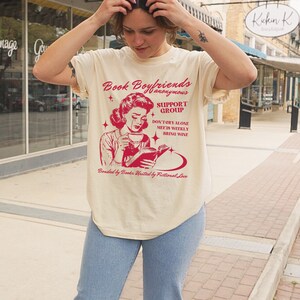 May include: A beige t-shirt with a red graphic of a woman reading a book and crying. The graphic says "Book Boyfriends Anonymous Support Group Don't Cry Alone Meets Weekly Bring Wine Bonded by Books United by Fictional Love."
