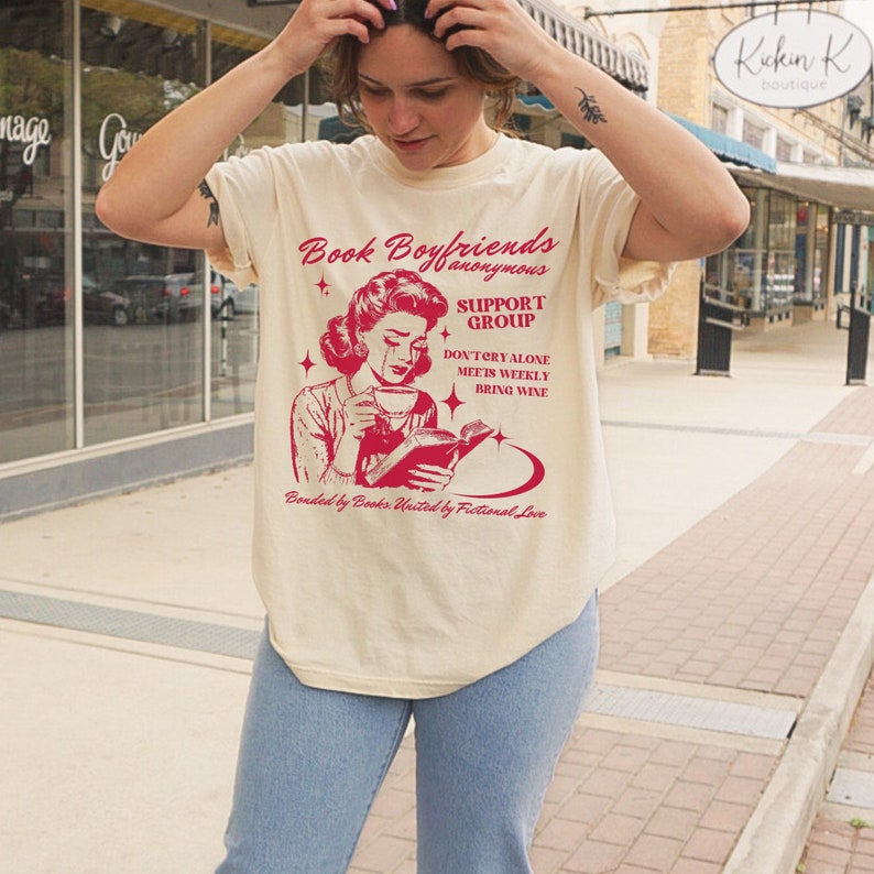 May include: A beige t-shirt with a red graphic of a woman reading a book and crying. The graphic says "Book Boyfriends Anonymous Support Group Don't Cry Alone Meets Weekly Bring Wine Bonded by Books United by Fictional Love."