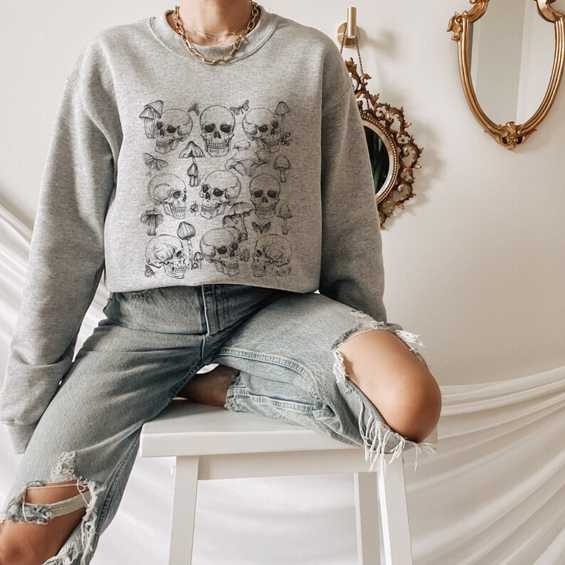 May include: A gray sweatshirt with a print of skulls and mushrooms. The sweatshirt has a crew neck and long sleeves.