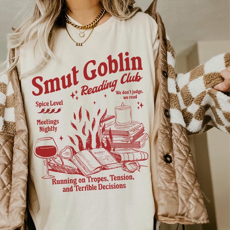 May include: A white t-shirt featuring a red graphic design that reads "Smut Goblin Reading Club". The graphic also includes a stack of books, a candle, and a glass of wine. The text below the graphic says "We don't judge, we read" and "Running on Tropes, Tension, and Terrible Decisions".