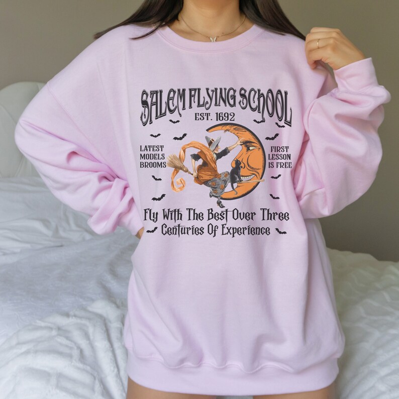 Op de afbeelding: Roze sweatshirt met een afbeelding van een heks die op een bezem vliegt met een zwarte kat. De afbeelding zegt "Salem Flying School Est. 1692" en "Fly With The Best Over Three Centuries Of Experience".