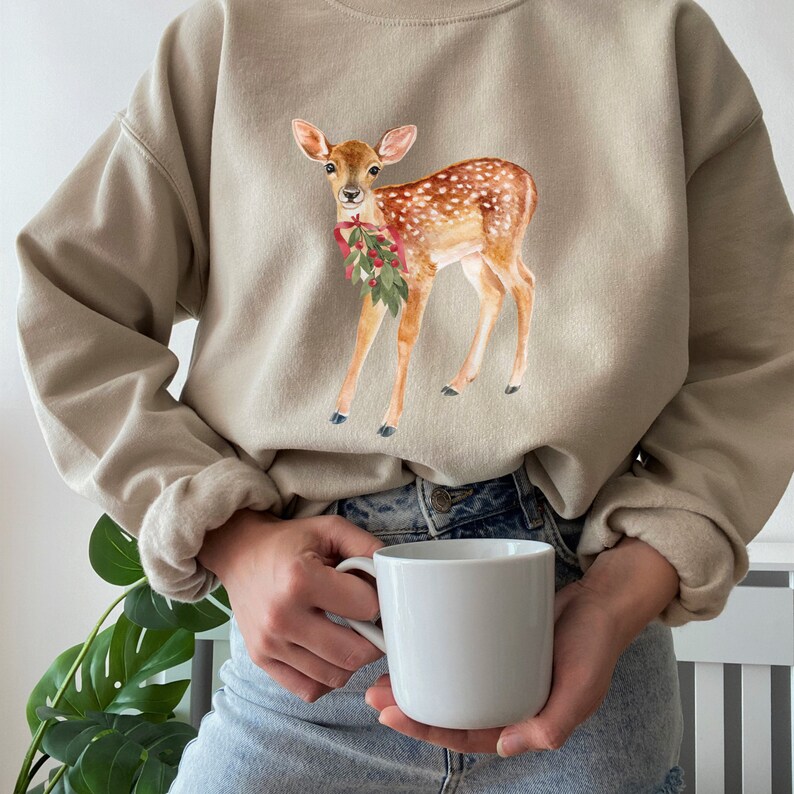 May include: A beige sweatshirt with a watercolor illustration of a fawn with a red bow and green leaves. The fawn is standing on all four legs and looking to the left.